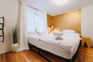 A hotel room with a bed, a bedside table, a lamp, a plant, and a window with curtains.