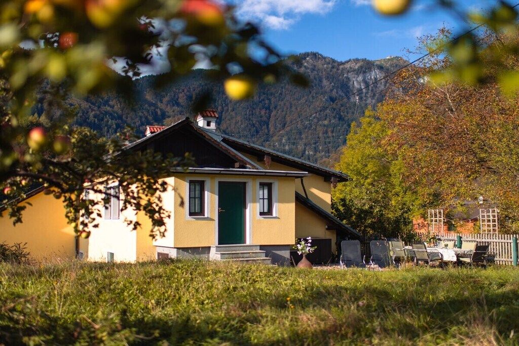 A yellow building with a green door, surrounded by grass and trees, with mountains in the background.