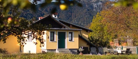 A yellow building with a green door, surrounded by grass and trees, with mountains in the background.