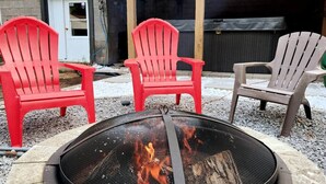 Uma fogueira com três cadeiras Adirondack, uma vermelha e duas cinzentas, em um quintal.