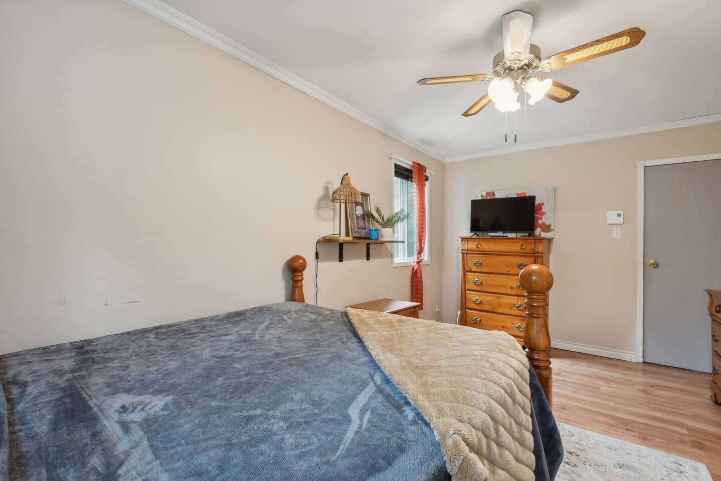 Une chambre à coucher avec un lit, une commode, un téléviseur posé sur le dessus, un ventilateur de plafond et une fenêtre avec des rideaux.