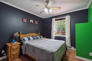 Un dormitorio con cama, mesita de noche, lámpara, ventilador de techo y ventana con vistas al verdor.