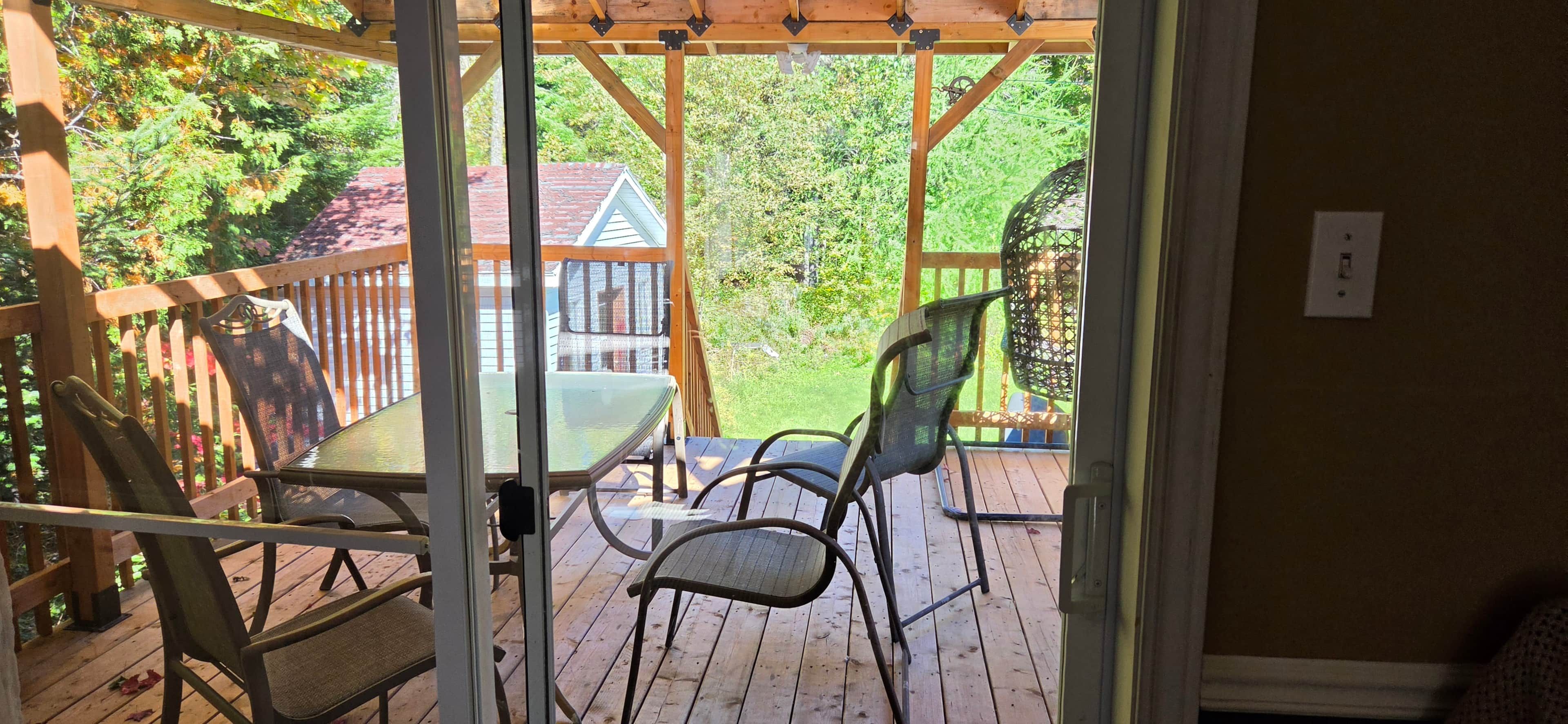Een overdekt terras met een tafel en stoelen, een houten terras en uitzicht op een woning en groen.