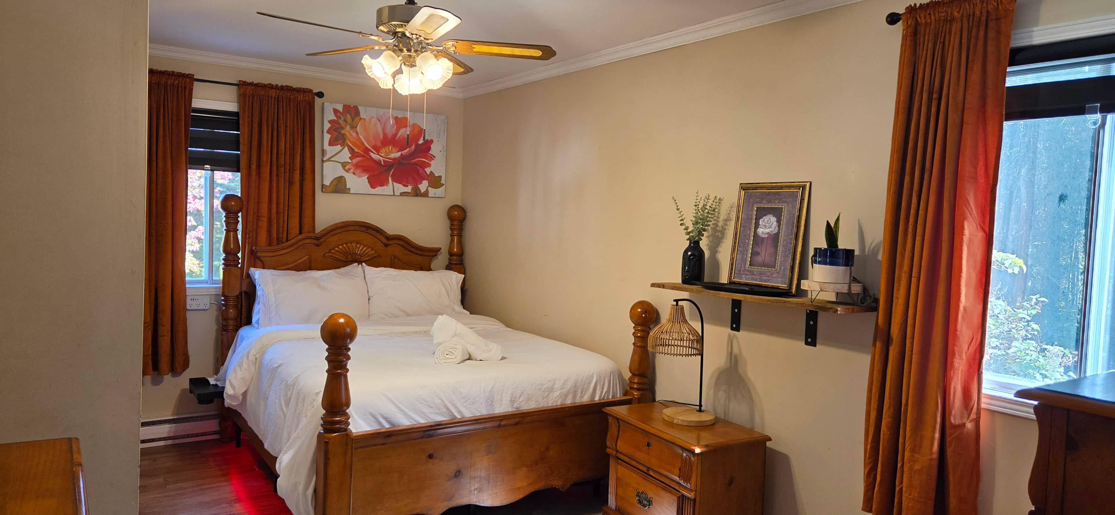 Ein Schlafzimmer mit einem hölzernen Bett, einem Nachttisch, einem Deckenventilator, einem Fenster mit Vorhängen und einer Wandablage mit Bilderrahmen und einer Vase.