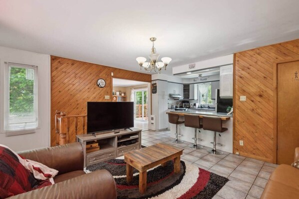Una sala de estar con televisor de pantalla plana, una mesa de centro de madera y sofás de cuero. Se ve una cocina con barra a través de un umbral abierto.