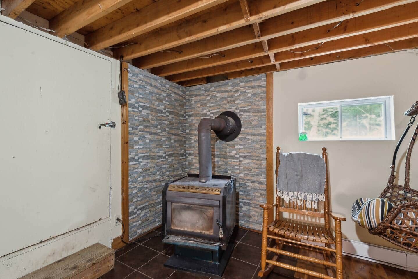 Une pièce avec un mur en pierre, un plafond en bois, un poêle rustique, une chaise en osier et une fenêtre donnant sur un paysage agréable.