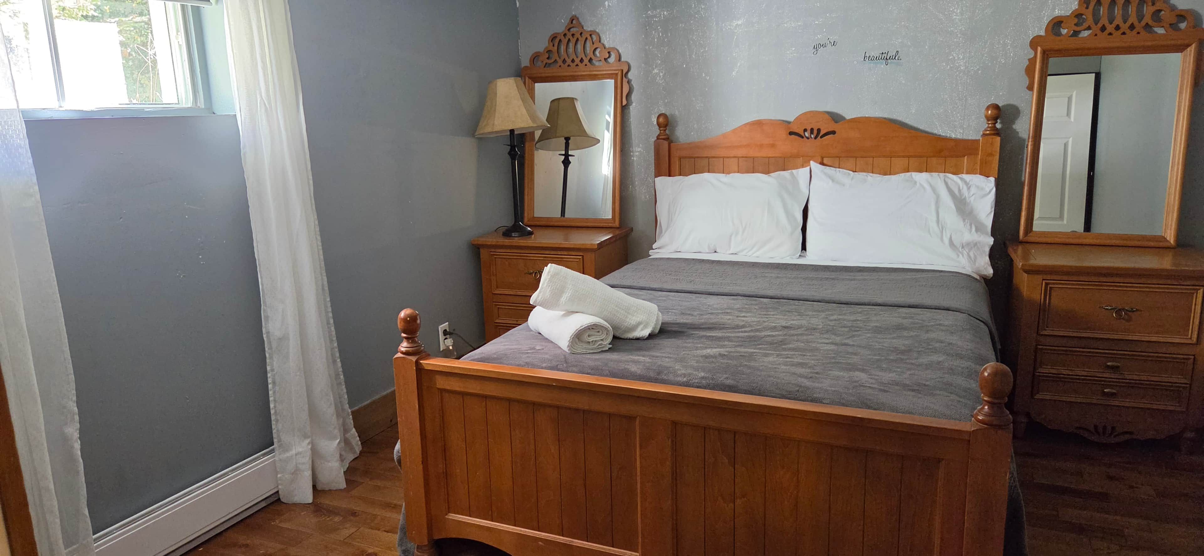 Un letto di legno con un materasso grigio, due comodini con lampade, uno specchio e una finestra con tende.