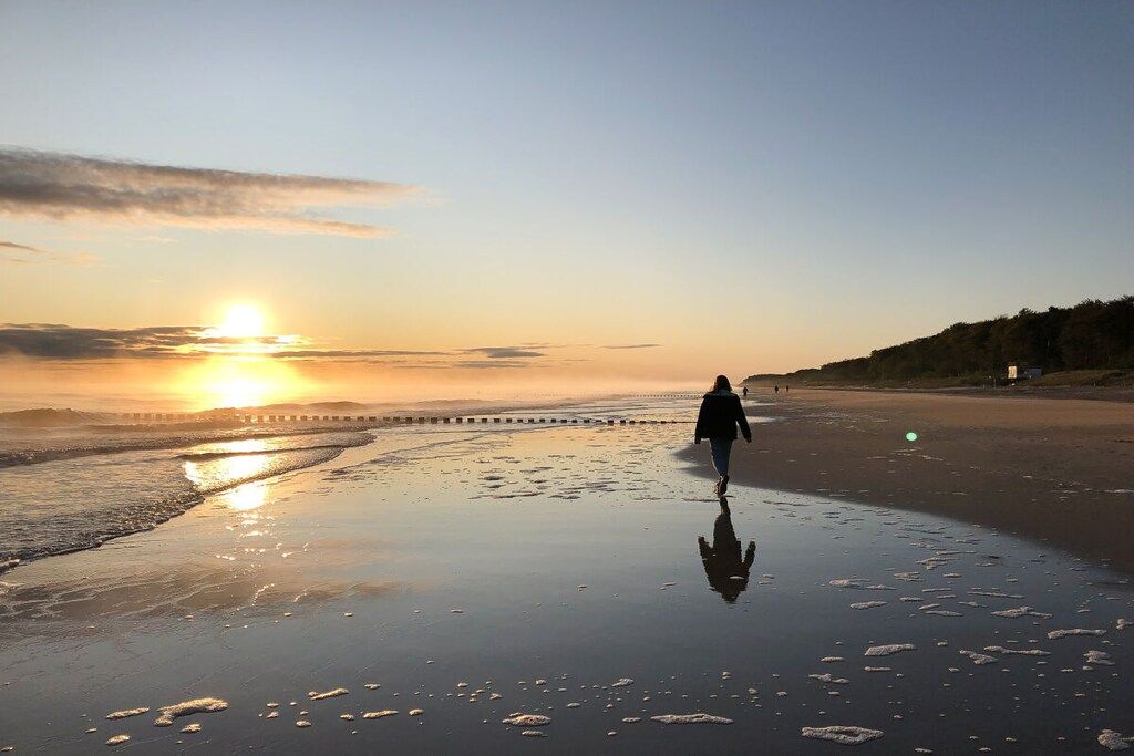 Une scène de plage où l’on voit une personne s’éloigner de la caméra, un coucher de soleil en arrière-plan et une mer calme.