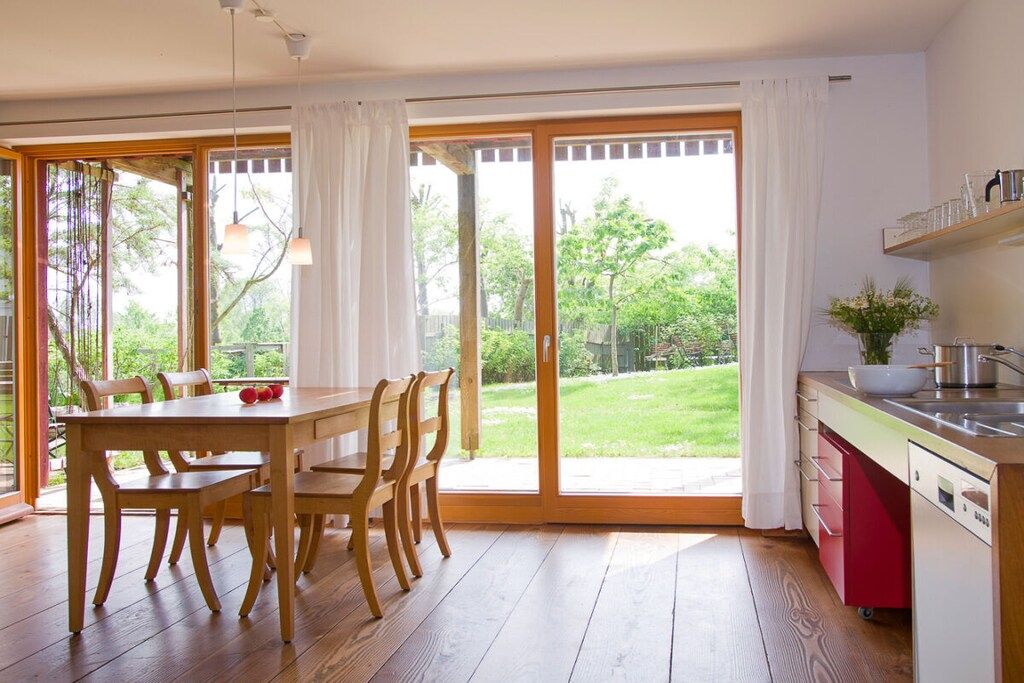 A room with a wooden dining table and chairs, a kitchen with a red cabinet, and large sliding glass doors leading to a garden.