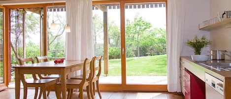 A room with a wooden dining table and chairs, a kitchen with a red cabinet, and large sliding glass doors leading to a garden.