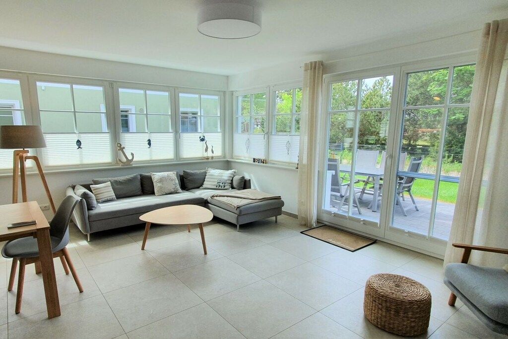 Un salon moderne avec un canapé gris, une table basse ronde et un coin repas comprenant une table et des chaises. De grandes fenêtres offrent une vue sur l’extérieur.