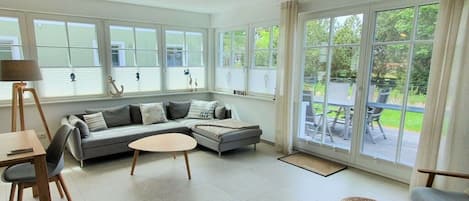 A modern living room with a grey sofa, a round coffee table, and a dining area with a table and chairs. Large windows provide a view of the outdoors.