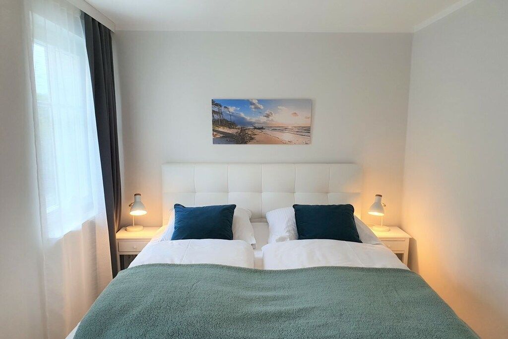 A hotel room with a bed, two bedside tables, a nightstand, a painting of a beach scene, and a window with curtains.
