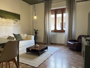 A living room with a sofa, a coffee table, a chair, a lamp, a radiator, and a window with curtains. - Bonaria: Apartment for 6 | Fiera, City Center, and Poetto (Cagliari)