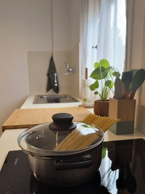 A kitchen with a pot on an induction cooktop, a wooden cutting board, a plant, and kitchen utensils. - Bonaria: Apartment for 6 | Fiera, City Center, and Poetto (Cagliari)