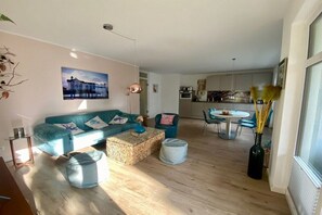 A living room with a sofa, coffee table, and ottoman, a dining area with a table and chairs, and a kitchen in the background. - Villa Louisa 43 - Octopussy (Ostseebad Sellin)