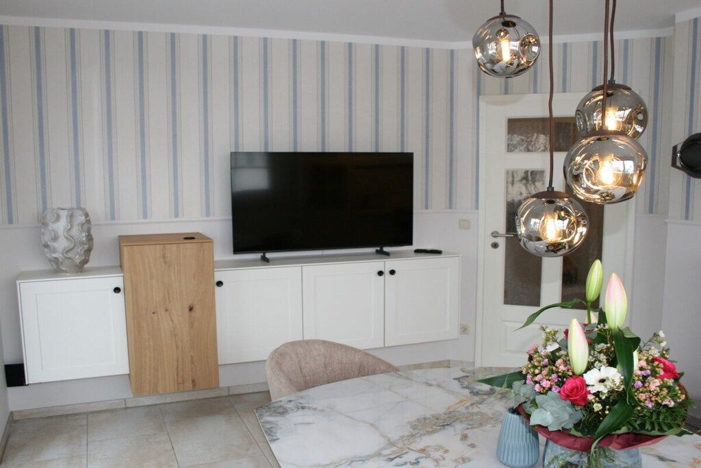 Uma sala de estar moderna com televisão de tela plana, armários brancos, aparador de madeira, mesa de jantar com padrão marmorizado e um arranjo floral.