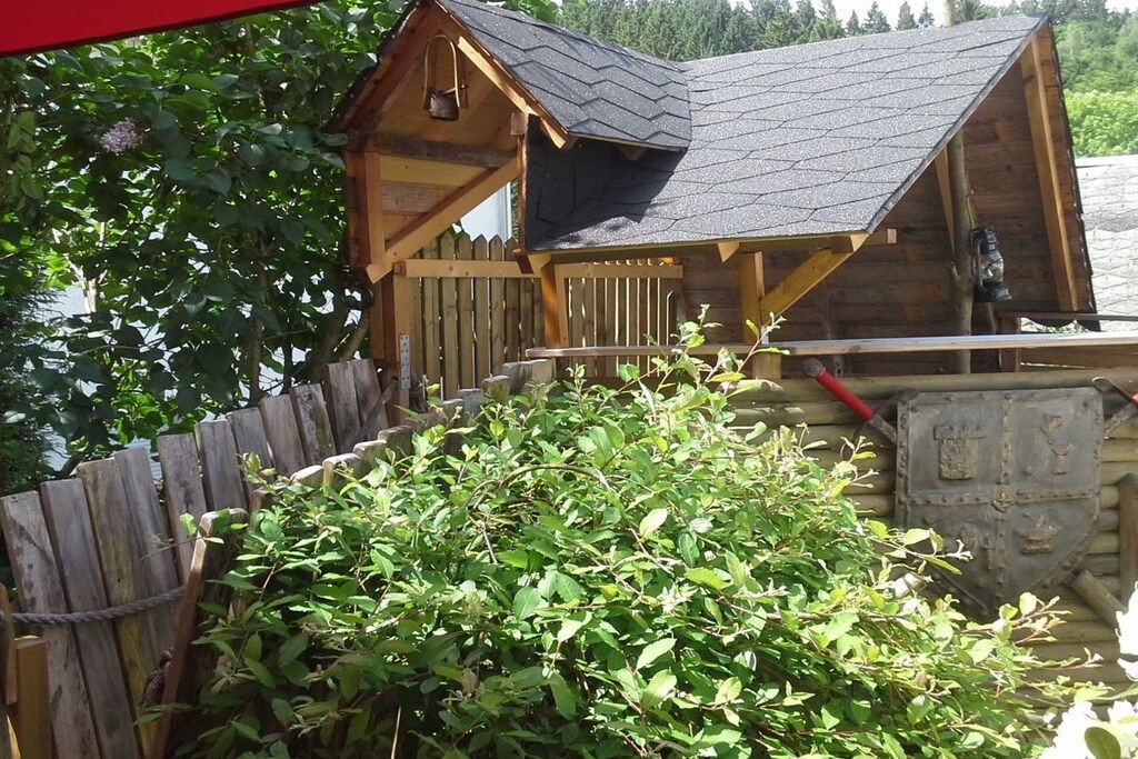 A wooden cabin with a sloped roof, surrounded by a wooden fence and dense foliage.