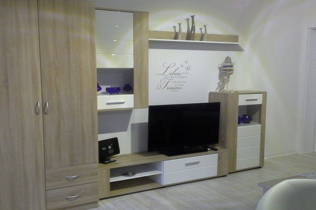 A room with a TV on a wooden stand, a cabinet with drawers, and a shelf with decorative items.