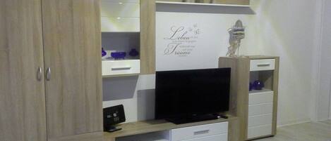 A room with a TV on a wooden stand, a cabinet with drawers, and a shelf with decorative items.