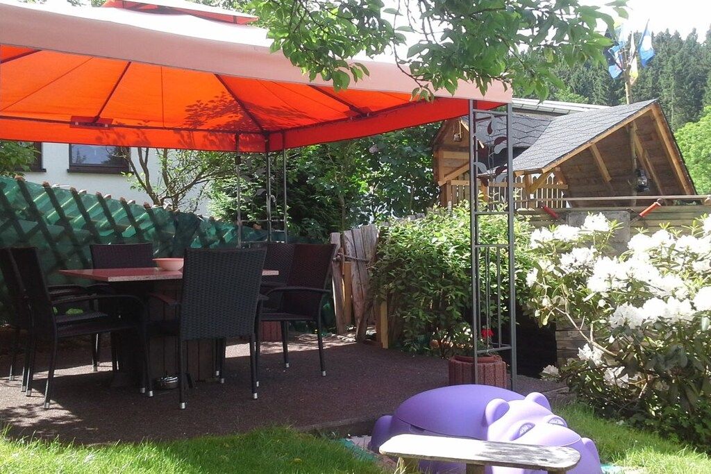 Un espace extérieur aménagé en terrasse, avec une table et des chaises sous un grand parasol, entouré de verdure et d’une structure en bois.