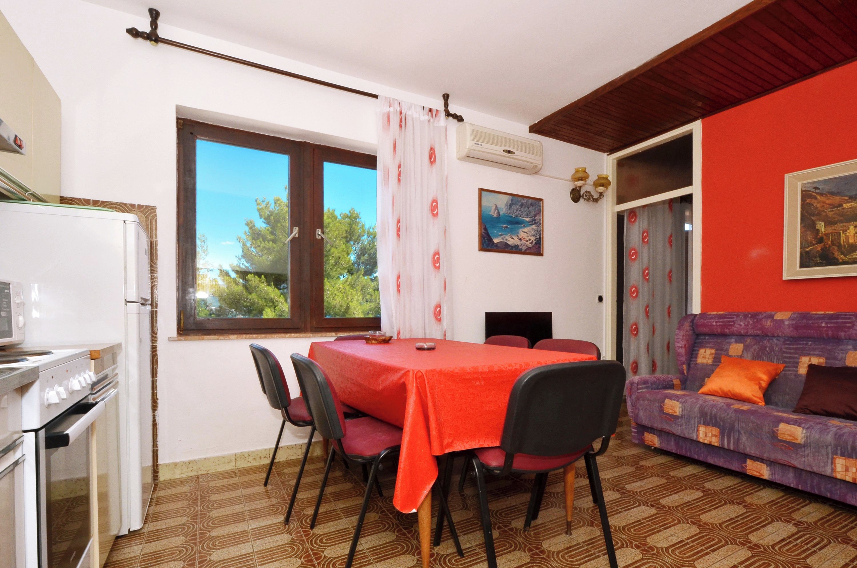 Ein Essbereich mit einem Tisch, der mit einer roten Tischdecke bedeckt ist, umgeben von Stühlen. Im Hintergrund befindet sich eine Couch mit Kissen und ein Fenster mit Vorhängen.
