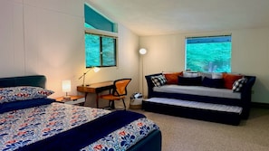 A bedroom with two beds, a desk, a chair, and two windows with a view of trees.
