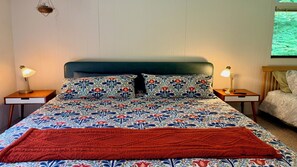Een bed met een bloemenbedekking, twee bedlampjes, een rode plaid en een raam met uitzicht op groen.