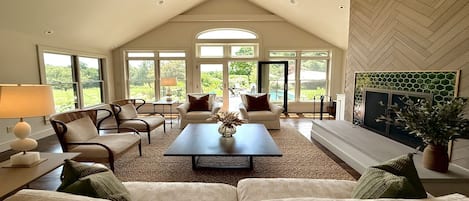 A spacious living room with a high ceiling, large windows, and a fireplace.