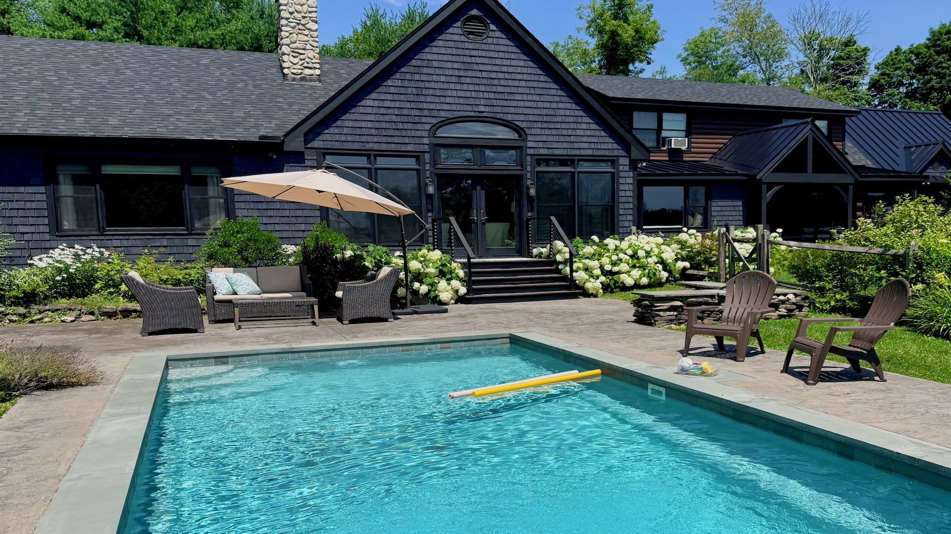 Una piscina con zona de estar, mobiliario exterior y una casa de fachada oscura y chimenea de piedra.
