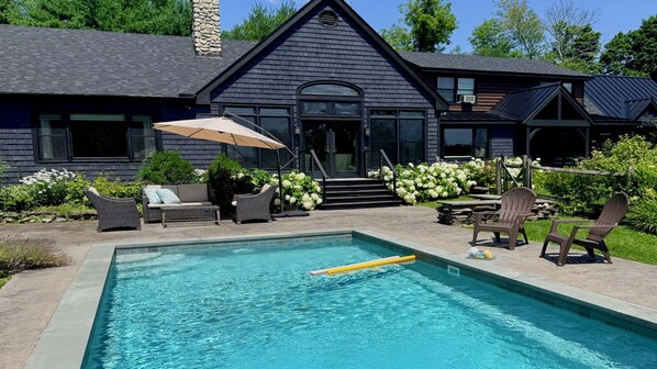 A swimming pool with a lounge area, outdoor furniture, and a house with a dark exterior and stone chimney.