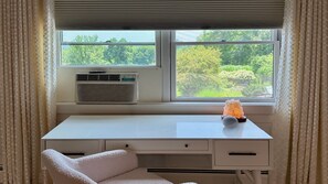 A room with a desk, a chair, a window with a view of greenery, and an air conditioning unit.