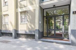 Entrada de um prédio com porta de vidro, placa com o número 5 e uma pequena área de estar ao ar livre.