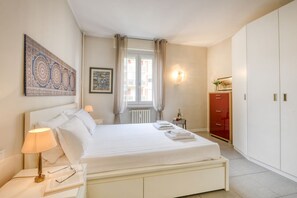 Quarto de hotel com uma cama grande, uma mesa de cabeceira com abajur, um guarda-roupa vermelho e uma janela com cortinas.