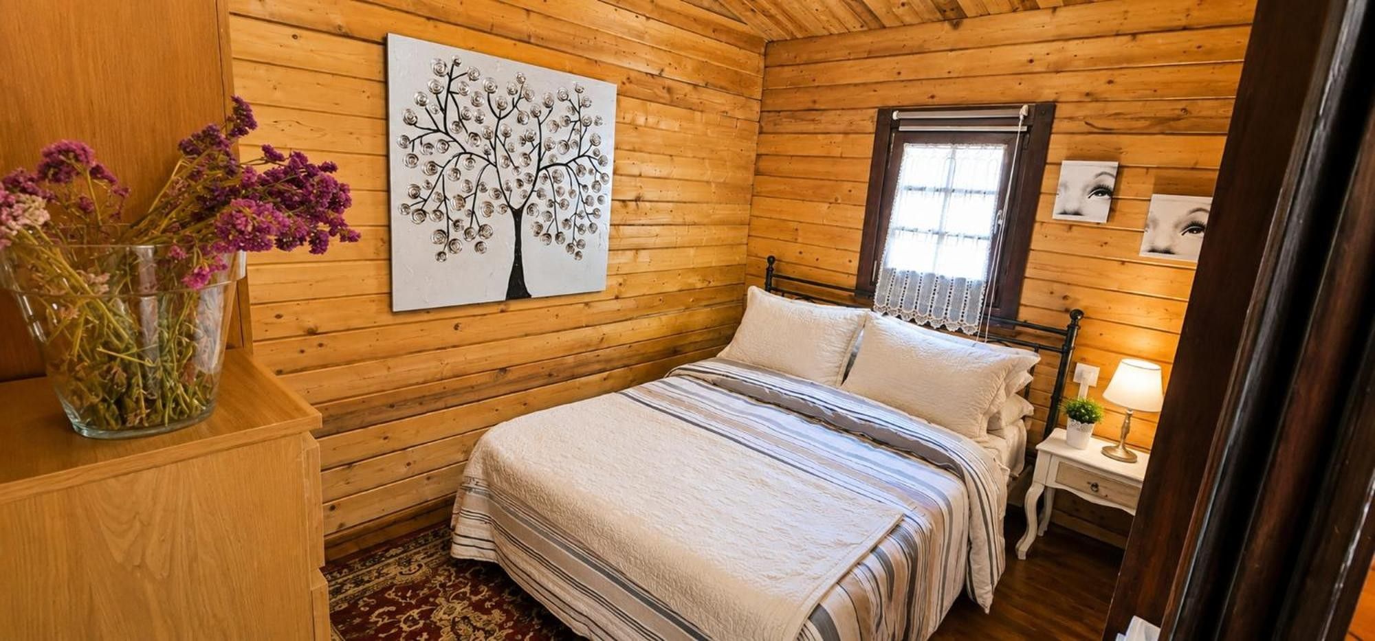 A wooden room with a bed, a wall-mounted picture, a vase with flowers, and a window.