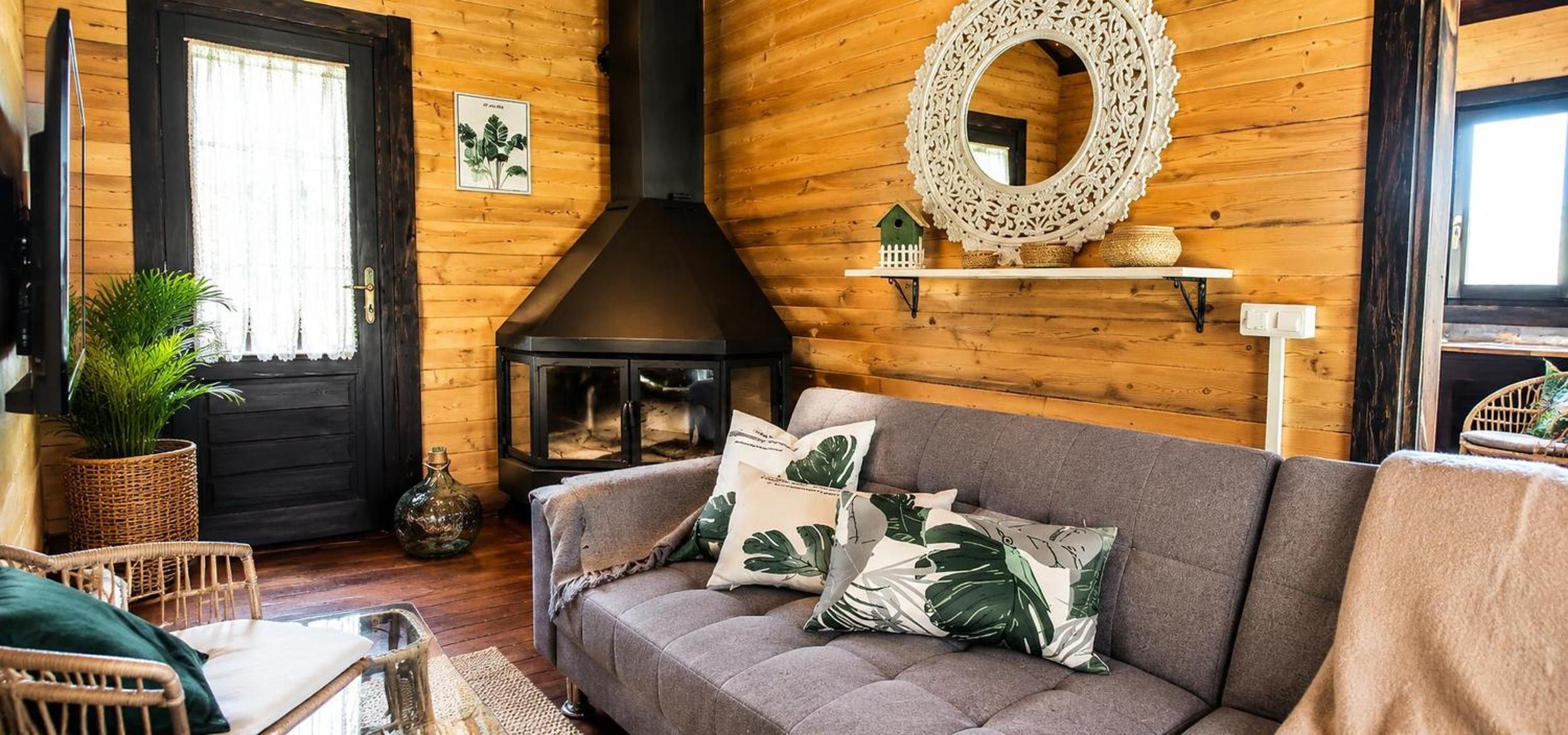 A cozy living room with a fireplace, a sofa with decorative pillows, wicker furniture, and a wooden wall with a decorative mirror.