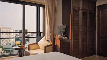 Una habitación de hotel con un ventanal grande, una cama, una silla, una mesita y un armario.