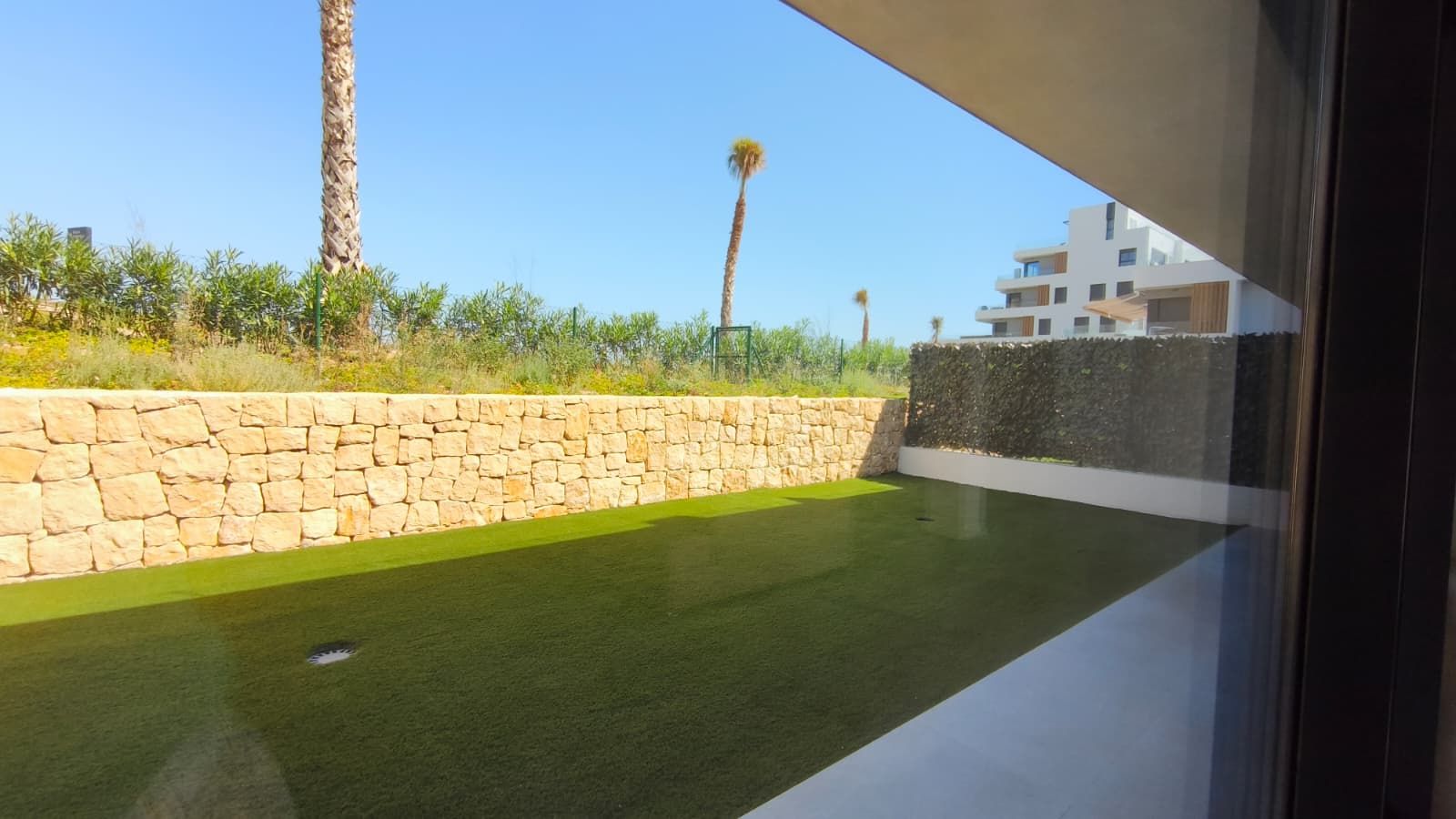 A pool with a stone wall and greenery, a clear sky, and a modern building in the background.