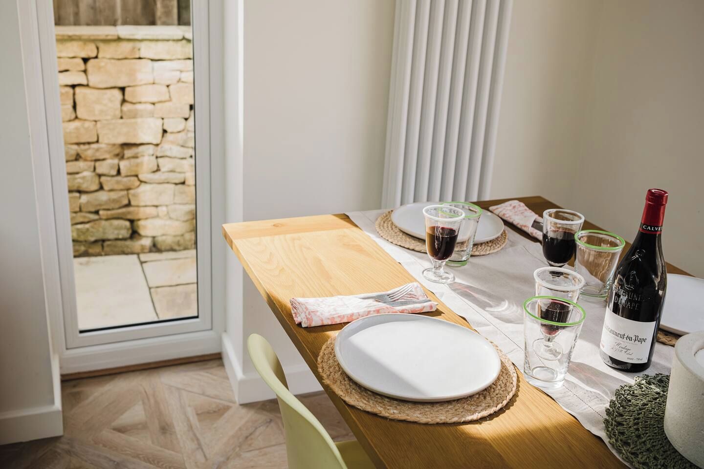 A dining table set with plates, glasses, and a bottle of wine, with a stone wall and a window in the background.