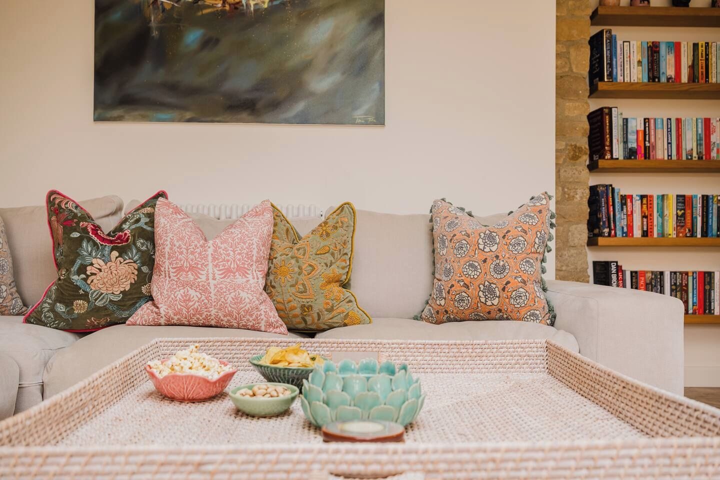 A living room with a sofa, decorative pillows, a woven tray with snacks, and a bookshelf filled with books.