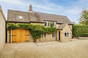 A two-story stone house with a wooden front door, a small window, and a gravel driveway. - Period Cotswold Cottage with outdoor bar & open views (Milton-under-Wychwood)