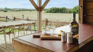 Puinen pöytä, jonka päällä on pullo Cotswolds Dry Gin -ginijuomaa, kaksi lasia ja kaksi Fever Tree -tonikavettä, ja pöytä on sijoitettu verannalle, jolta avautuu näkymä peltoalueelle.