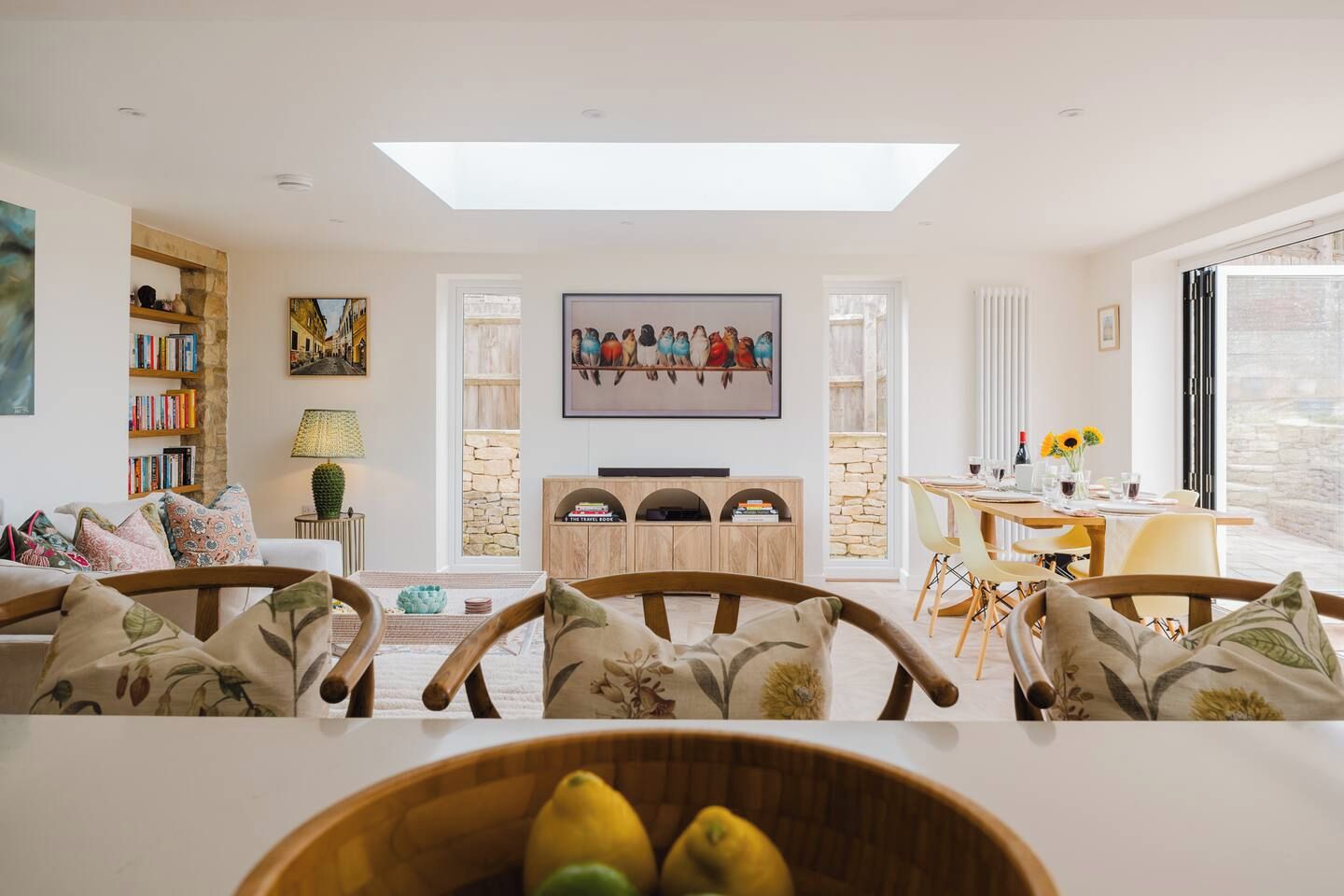 A modern living room with a high ceiling, a central fireplace, a dining area with a table and chairs, and a bookshelf filled with books.