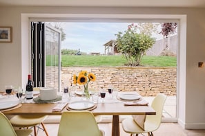 An open dining area with a table set for four, a bottle of wine, and a vase with sunflowers. - Period Cotswold Cottage with outdoor bar & open views (Milton-under-Wychwood)