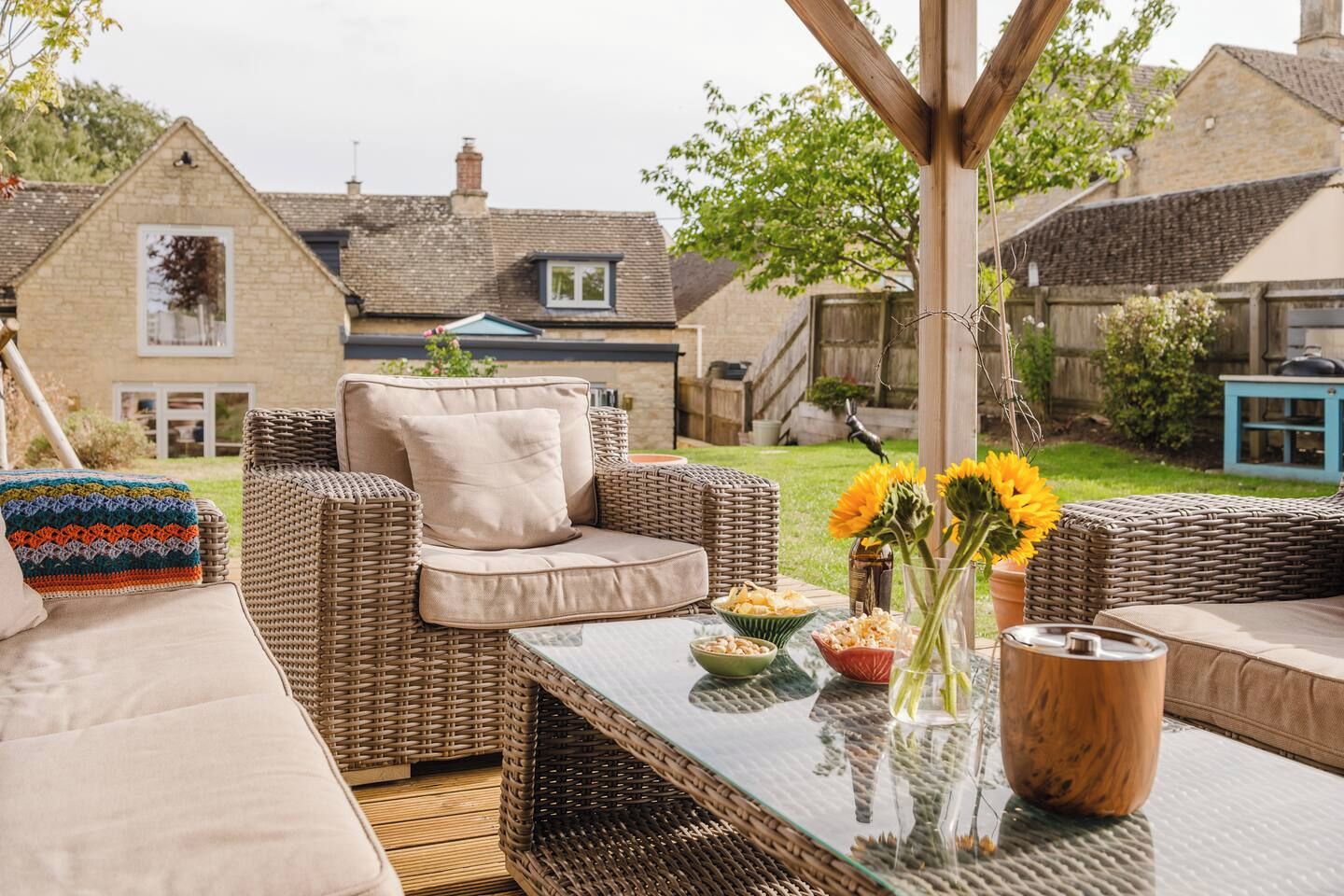 A wicker sofa set with cushions, a glass-top table with food and drinks, and a view of a house in the background.