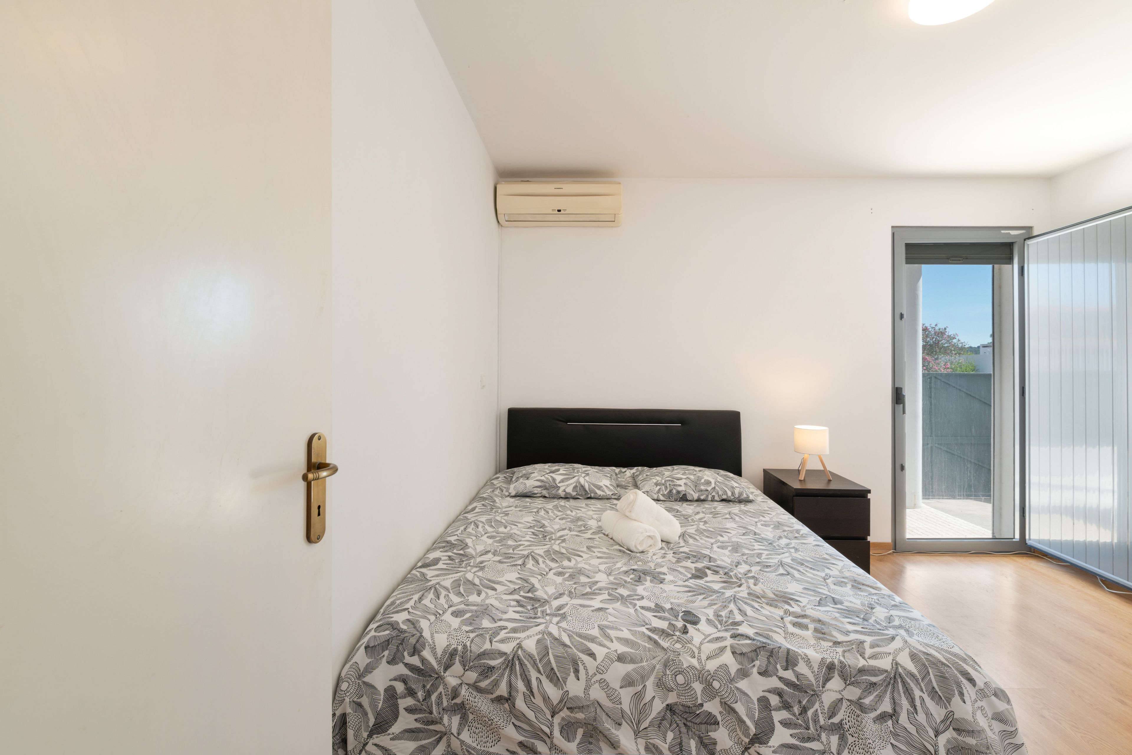 Ein Schlafzimmer mit einem Bett, einem Nachttisch, einer Lampe, einer Klimaanlage und einer Glastür, die zu einem Außenbereich führt.