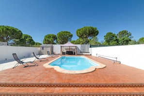 A swimming pool with lounge chairs and a covered seating area, surrounded by trees and a white wall.
