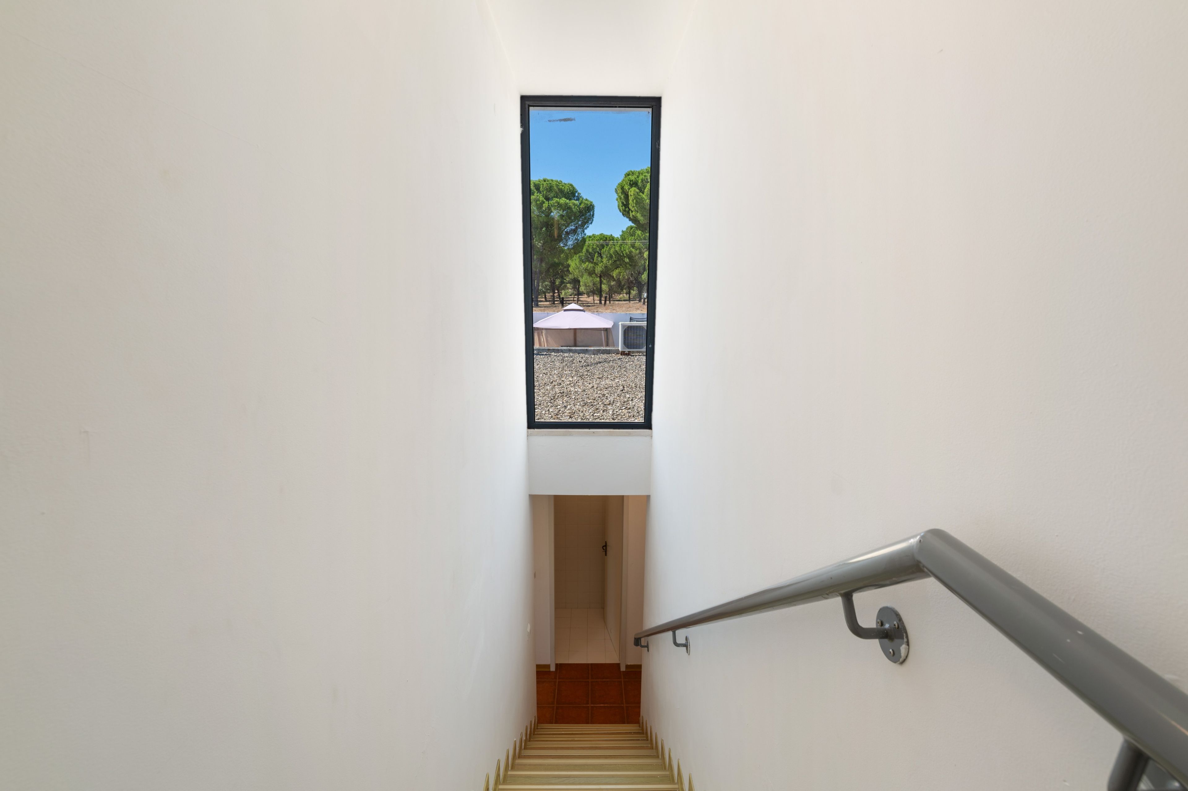 A narrow stairway leading to a landing with a small window overlooking a courtyard with trees and outdoor furniture.