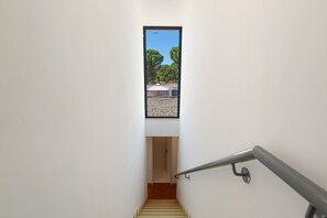 Un escalier étroit mène à un palier doté d’une petite fenêtre donnant sur une cour intérieure agrémentée d’arbres et de mobilier d’extérieur.