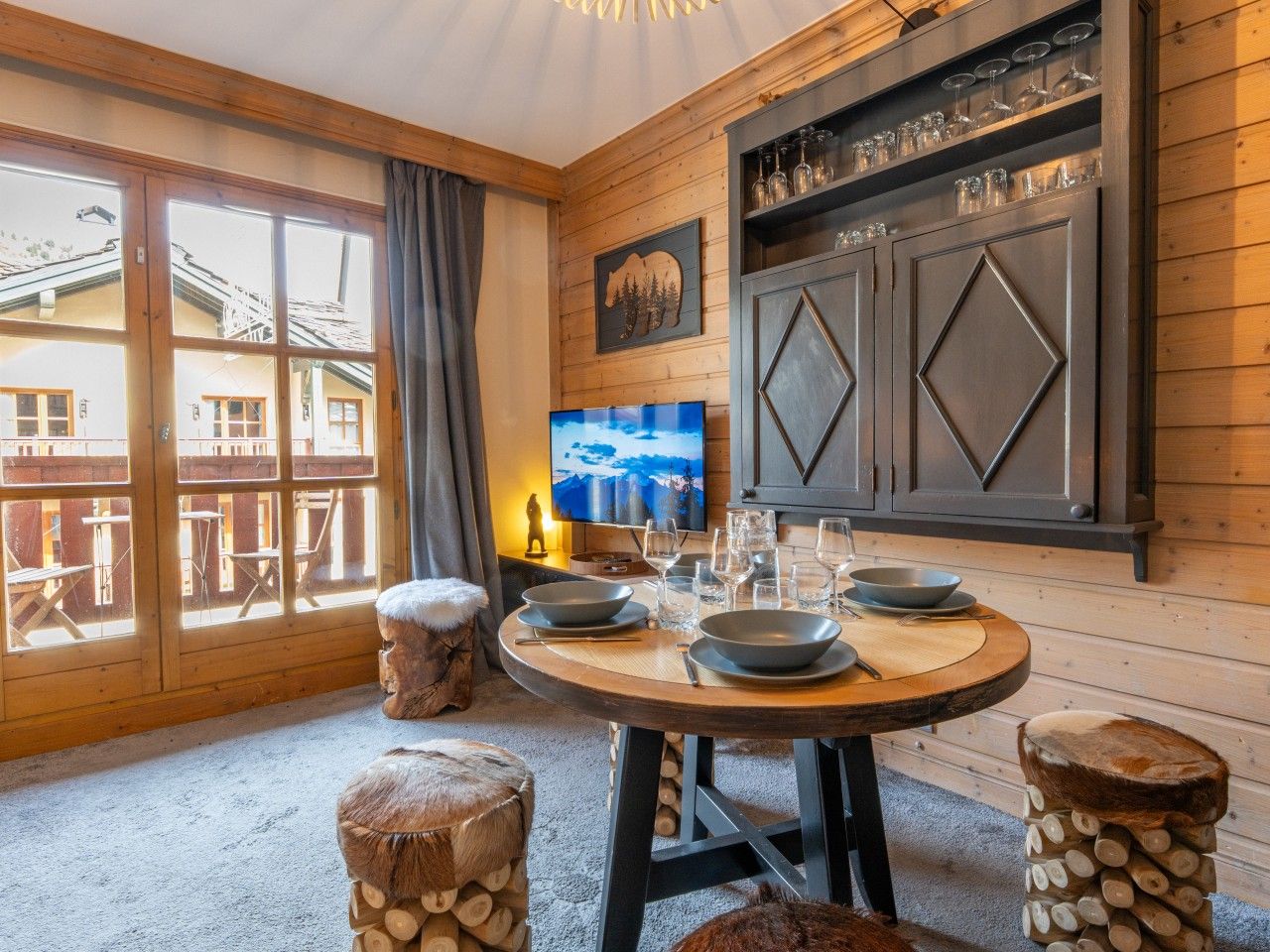 A dining area with a round table, wooden stools, and a television on the wall.